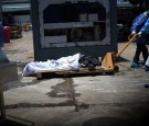 Health workers wearing protective gear bring a dead body past a refrigerated container outside of Teodoro Maldonado Carbo Hospital