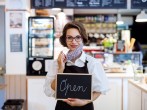 Young woman with face mask working in cafe, holding open sign. — Stock Image